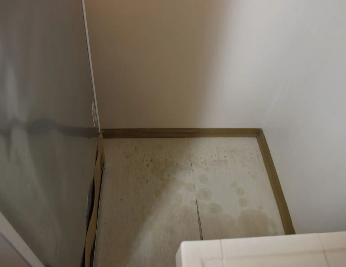 Pale floor behind a refrigerator with scattered marks and debris.