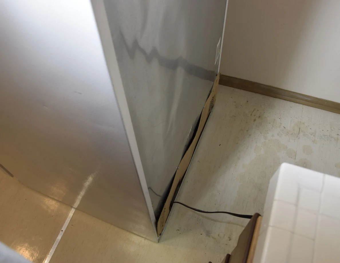 Lower rear of refrigerator with cord crossing marked pale flooring.