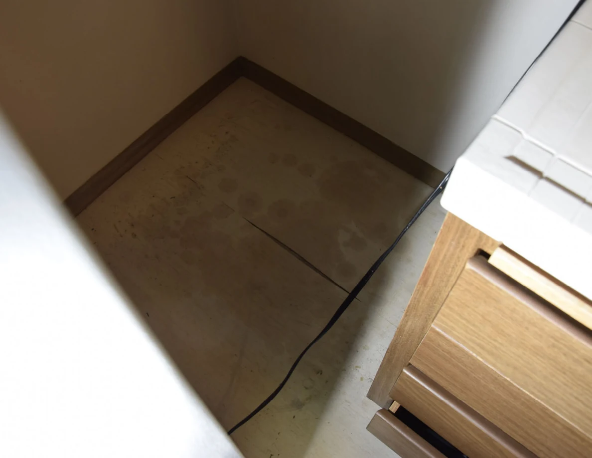 Floor corner behind refrigerator beside a wooden drawer unit and power cord.
