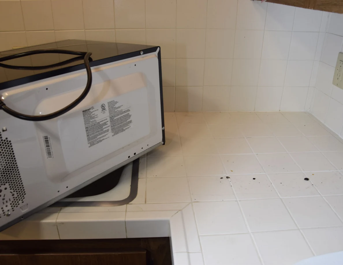 Underside of a microwave beside scattered dark debris on white counter tiles.