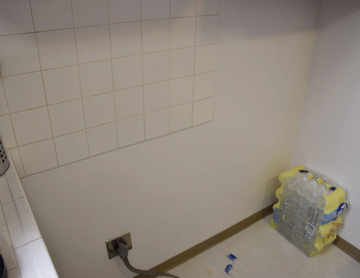 Tiled wall and outlet behind a moved stove, with packs of drinks below.