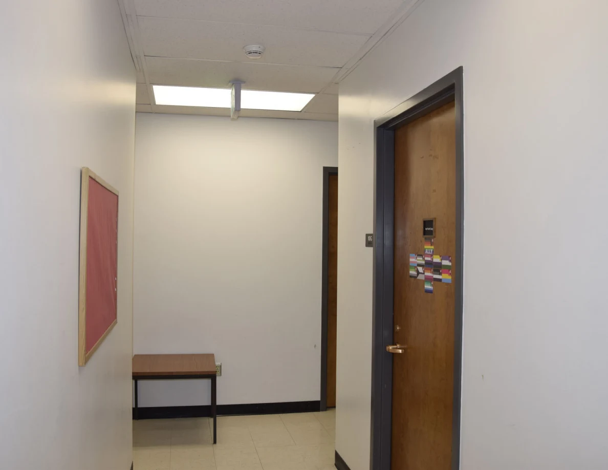 Office corridor with wooden doors, bulletin board, small table and ceiling lights.