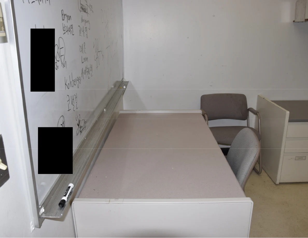 Desk and chairs below a whiteboard with writing partly covered by black redactions.