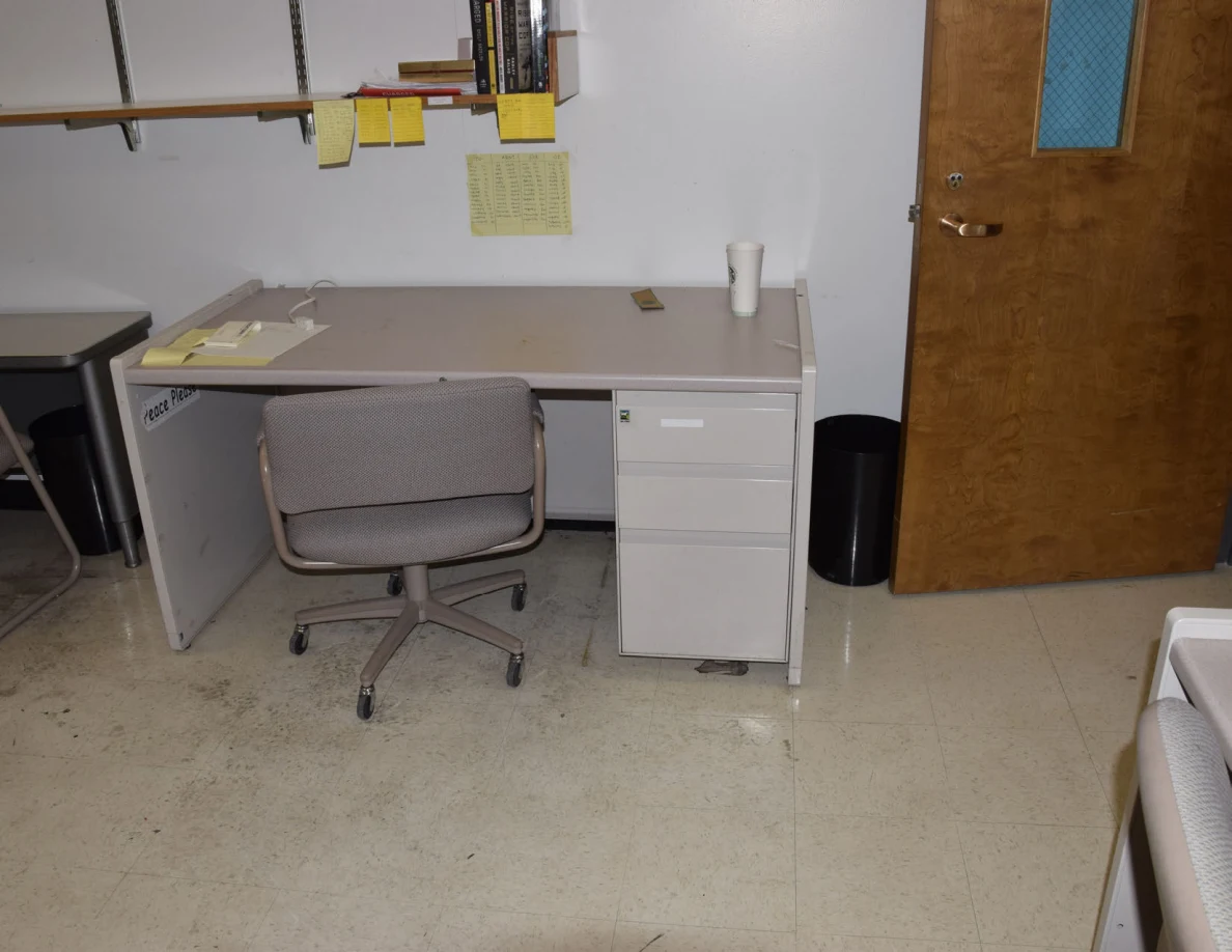 Office desk with rolling chair, telephone, white cup and small wastebasket beside a door.