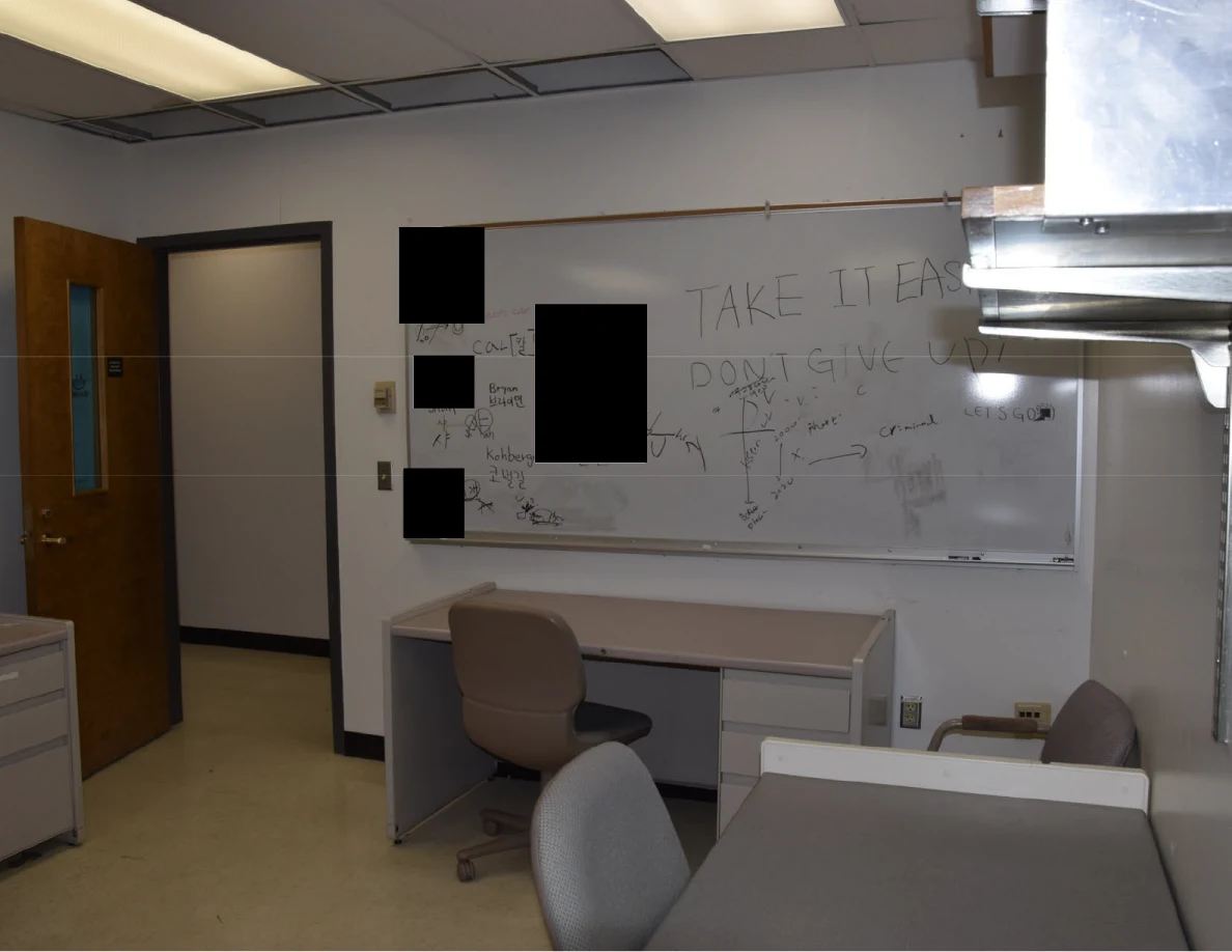 Whiteboard with handwritten messages and black redactions above a desk and chair.