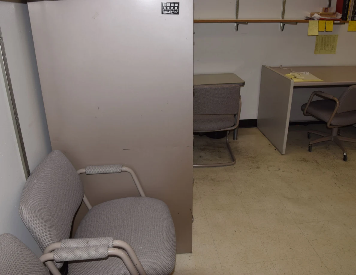 Upholstered visitor chair beside a tall filing cabinet with desks beyond.