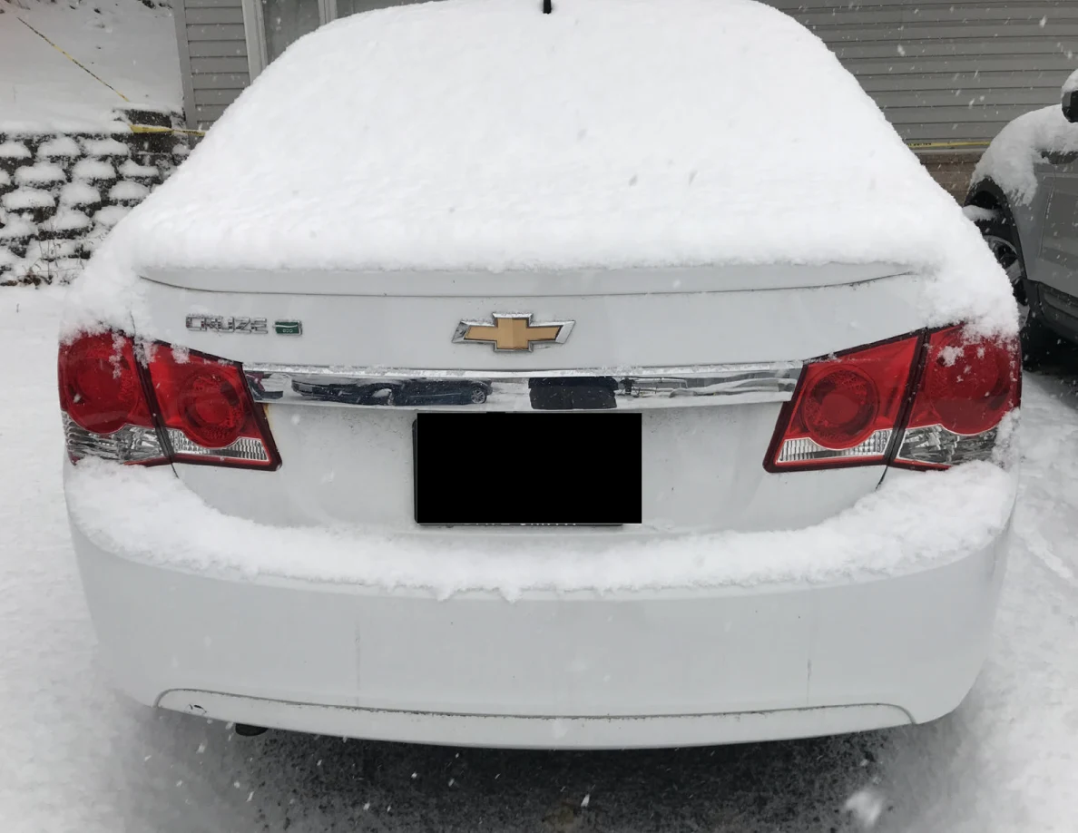 Snow-covered rear of a white Chevrolet Cruze with license plate blacked out.