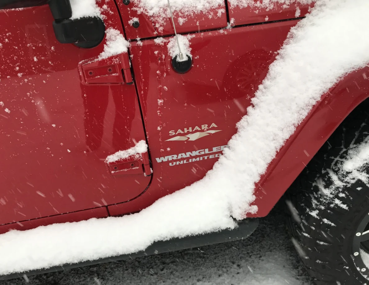 Snow-covered side of a red Jeep Wrangler Unlimited Sahara near its front wheel.