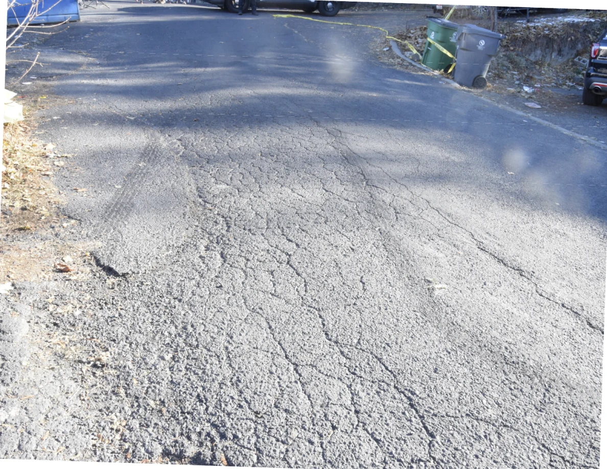 Cracked asphalt roadway with parked vehicles and wheeled bins in the distance.