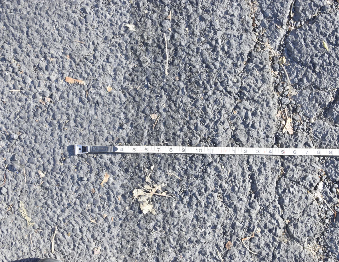 Close view of measuring tape beside rough asphalt and dry plant debris.