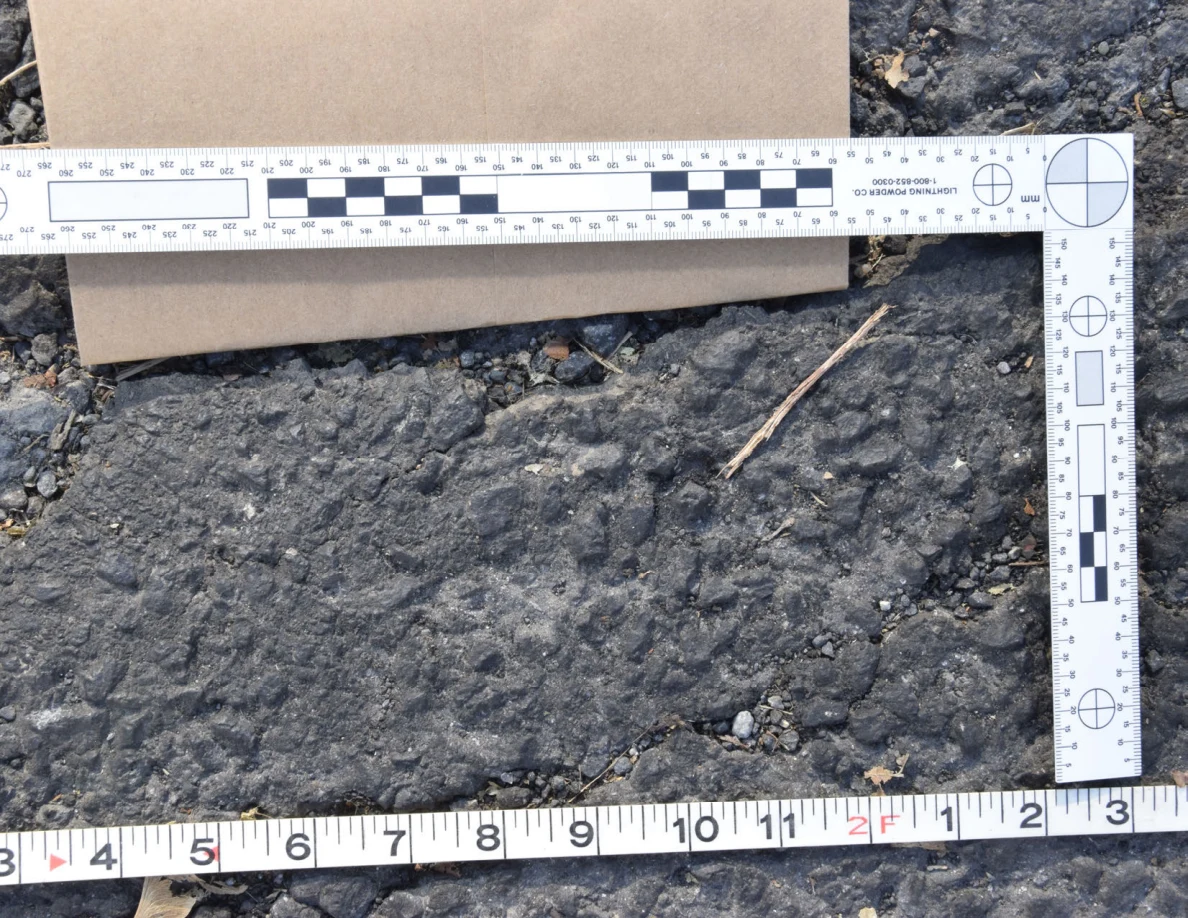Close view of a rough asphalt patch with a twig and small stones, framed by photographic scale and tape measure.