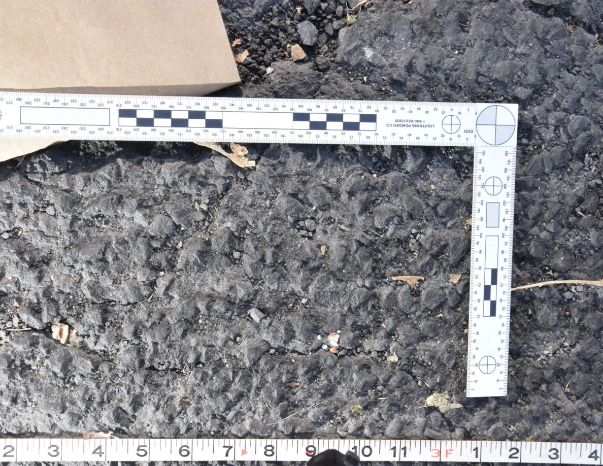 Textured asphalt with small pebbles and leaf fragments inside an L-shaped photographic scale, beside a brown envelope.