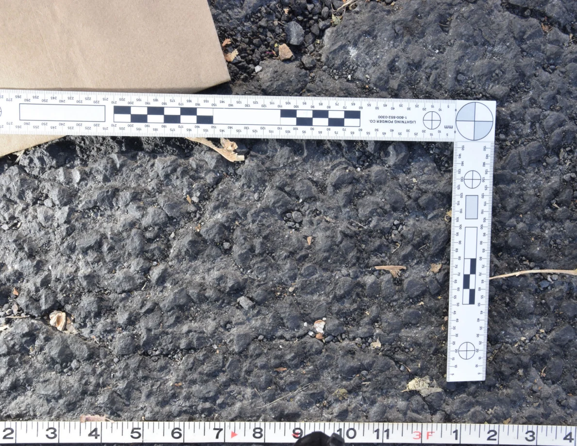 Textured asphalt with small pebbles and leaf fragments inside an L-shaped photographic scale, beside a brown envelope.