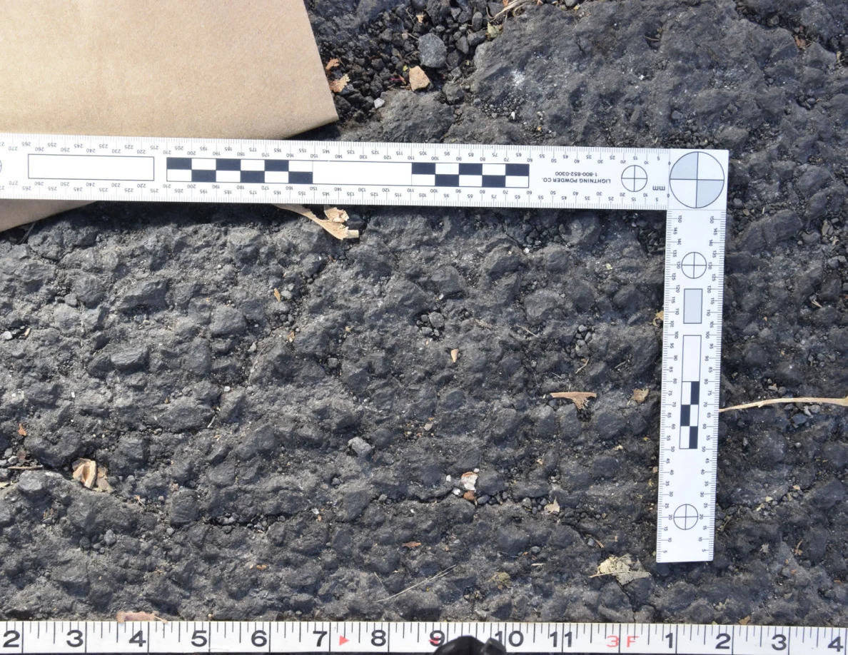 Textured asphalt with small pebbles and leaf fragments inside an L-shaped photographic scale, beside a brown envelope.