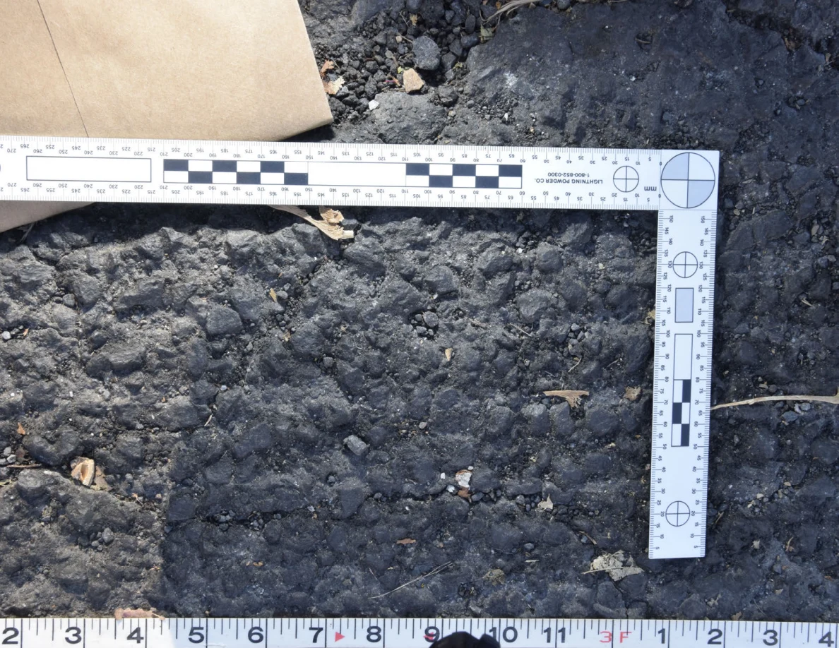 Textured asphalt with small pebbles and leaf fragments inside an L-shaped photographic scale, beside a brown envelope.