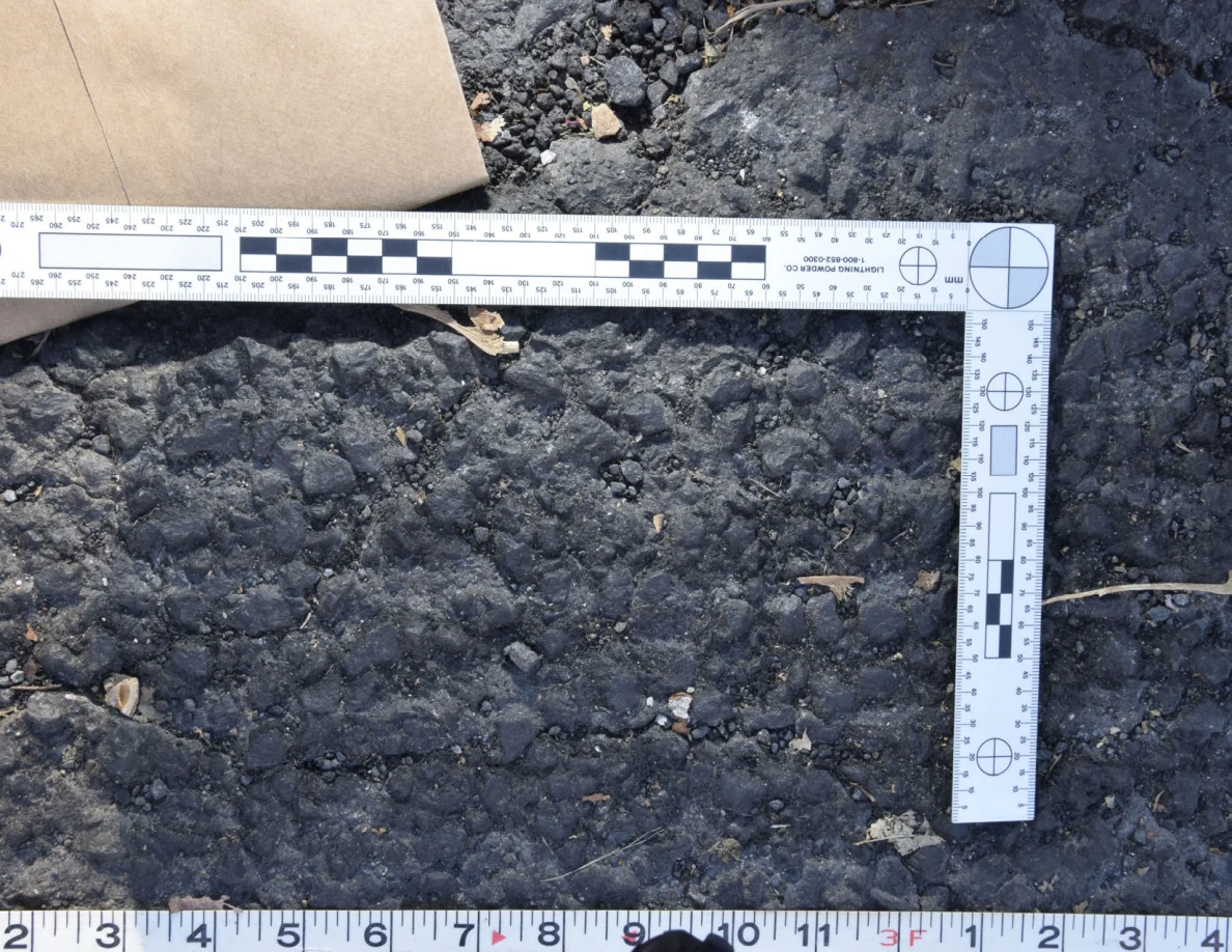 Textured asphalt with small pebbles and leaf fragments inside an L-shaped photographic scale, beside a brown envelope.