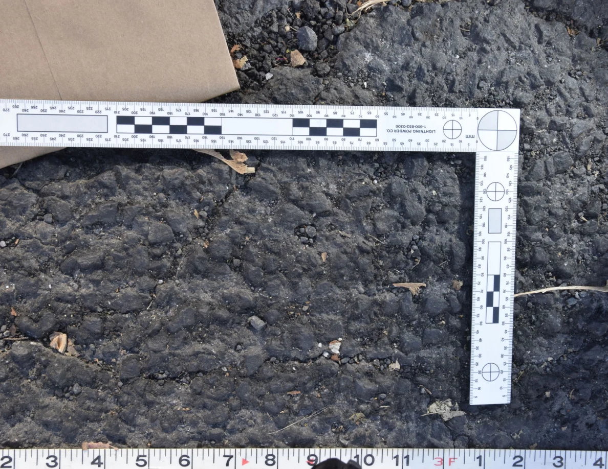 Textured asphalt with small pebbles and leaf fragments inside an L-shaped photographic scale, beside a brown envelope.