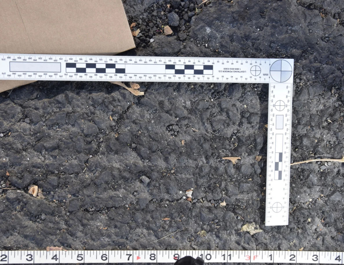 Textured asphalt with small pebbles and leaf fragments inside an L-shaped photographic scale, beside a brown envelope.
