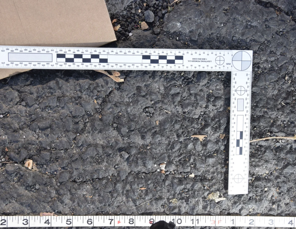 Textured asphalt with small pebbles and leaf fragments inside an L-shaped photographic scale, beside a brown envelope.