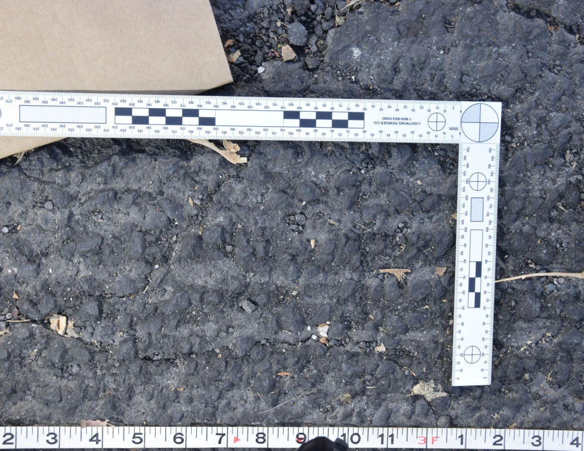 Textured asphalt with small pebbles and leaf fragments inside an L-shaped photographic scale, beside a brown envelope.