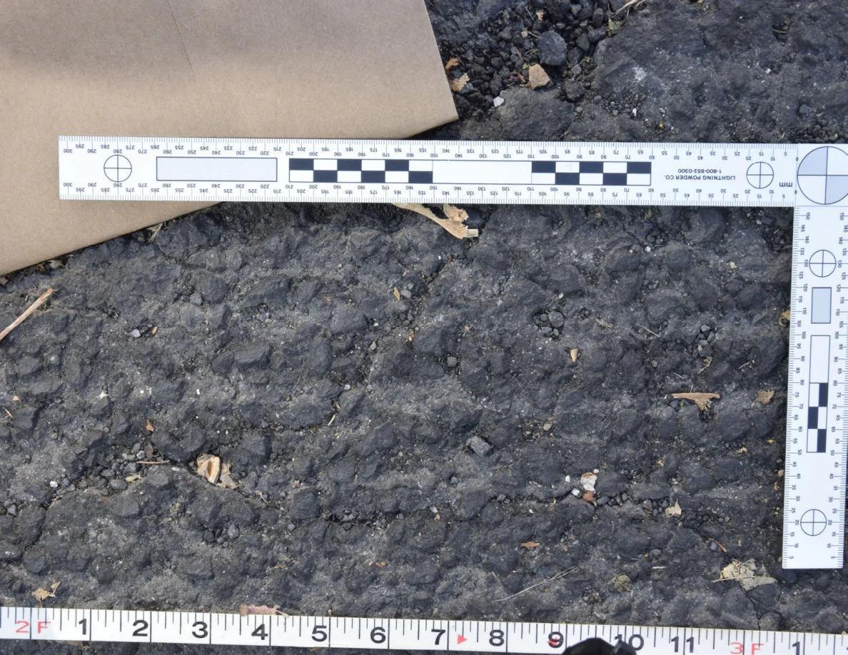 Close view of uneven roadway texture below a brown envelope and L-shaped scale, with a tape measure along the bottom.