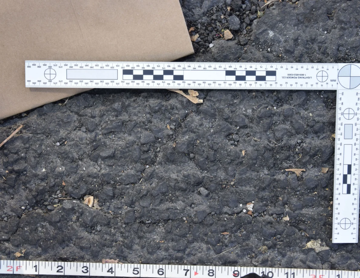 Close view of uneven roadway texture below a brown envelope and L-shaped scale, with a tape measure along the bottom.