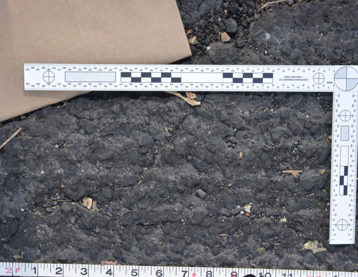 Close view of uneven roadway texture below a brown envelope and L-shaped scale, with a tape measure along the bottom.