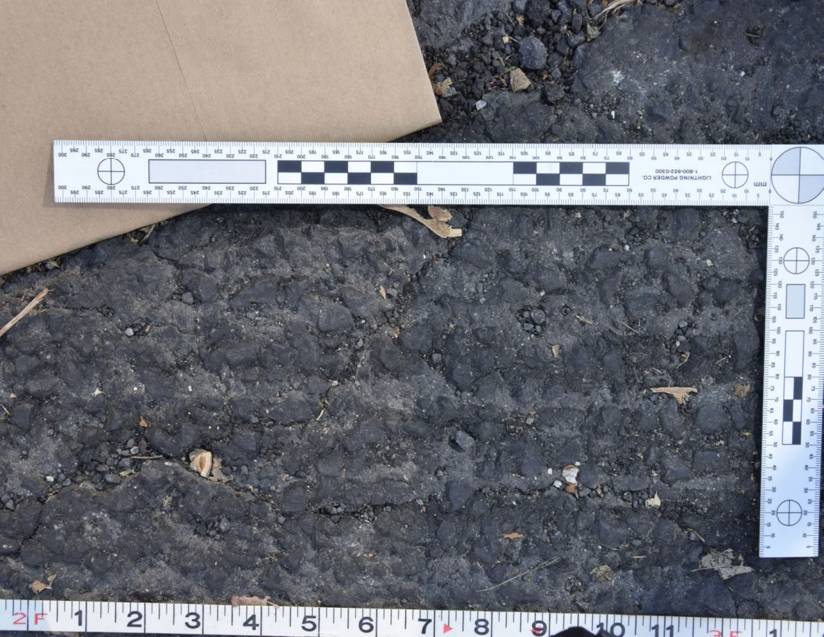 Close view of uneven roadway texture below a brown envelope and L-shaped scale, with a tape measure along the bottom.