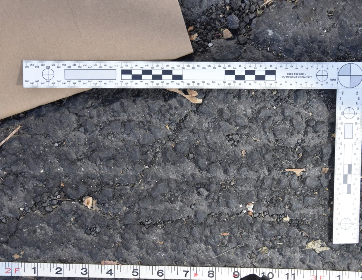 Close view of uneven roadway texture below a brown envelope and L-shaped scale, with a tape measure along the bottom.
