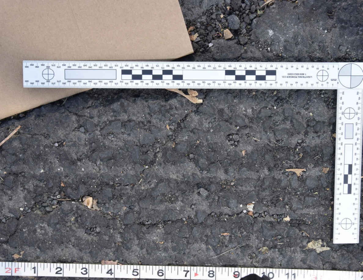 Close view of uneven roadway texture below a brown envelope and L-shaped scale, with a tape measure along the bottom.