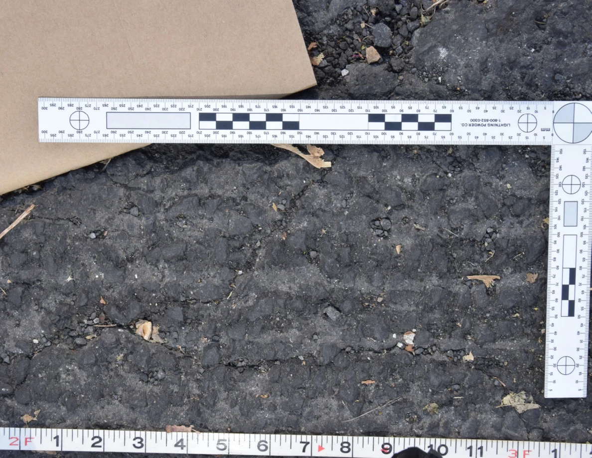 Close view of uneven roadway texture below a brown envelope and L-shaped scale, with a tape measure along the bottom.