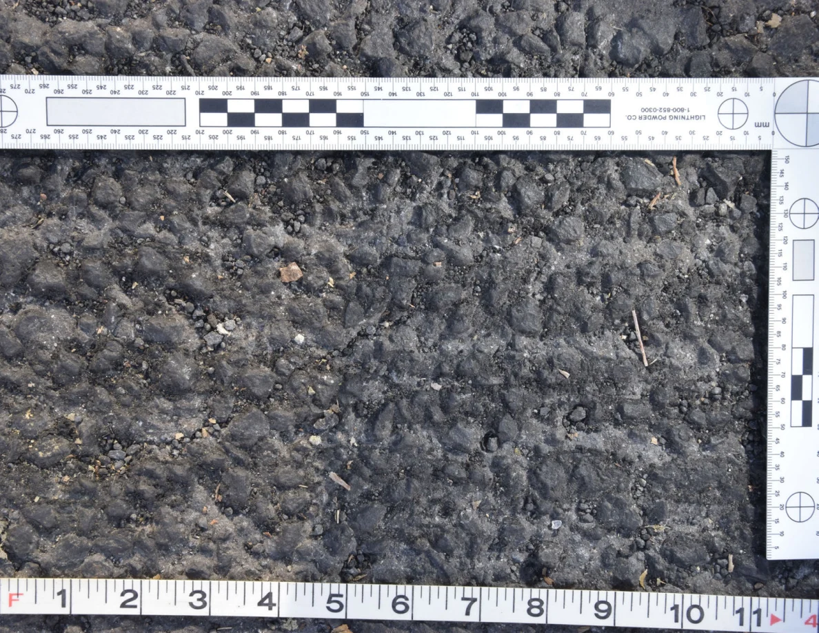 Close view of rough asphalt with small stones and thin plant fragments beside an L-shaped scale and measuring tape.