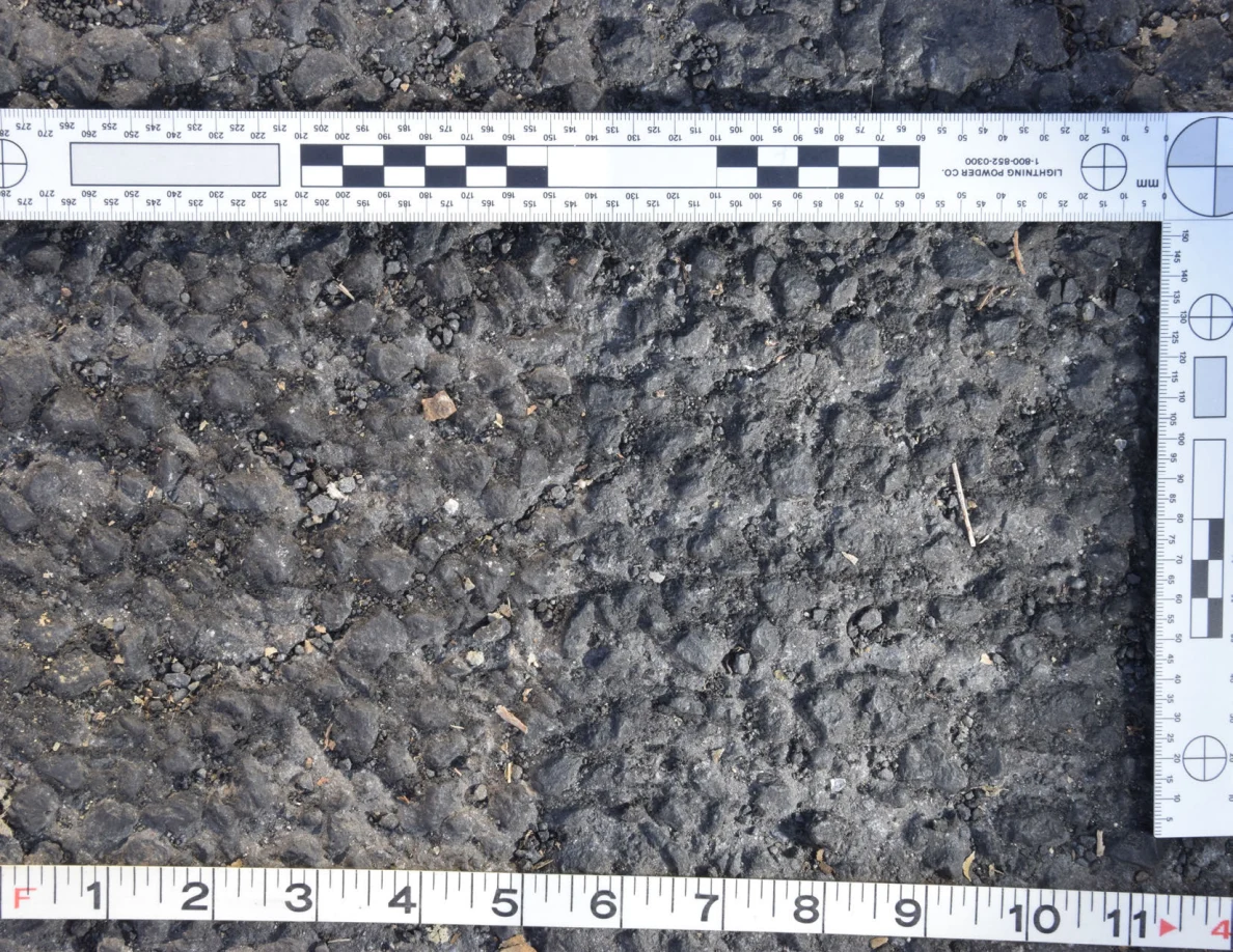 Close view of rough asphalt with small stones and thin plant fragments beside an L-shaped scale and measuring tape.