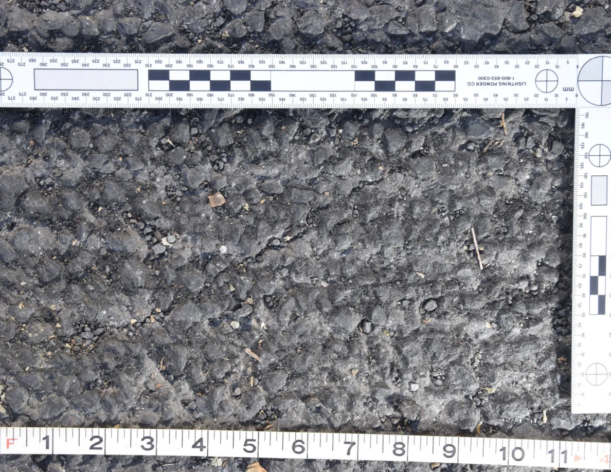 Close view of rough asphalt with small stones and thin plant fragments beside an L-shaped scale and measuring tape.