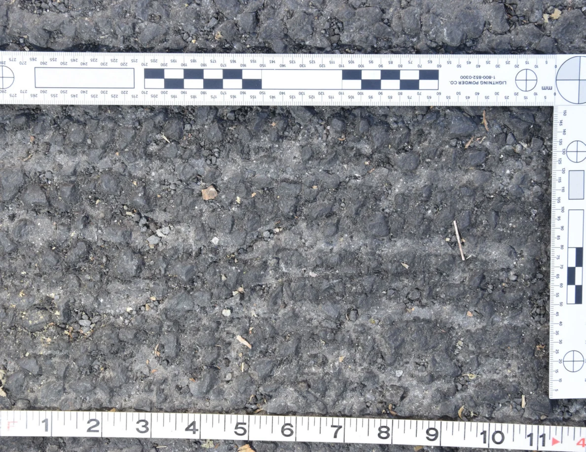 Close view of rough asphalt with small stones and thin plant fragments beside an L-shaped scale and measuring tape.