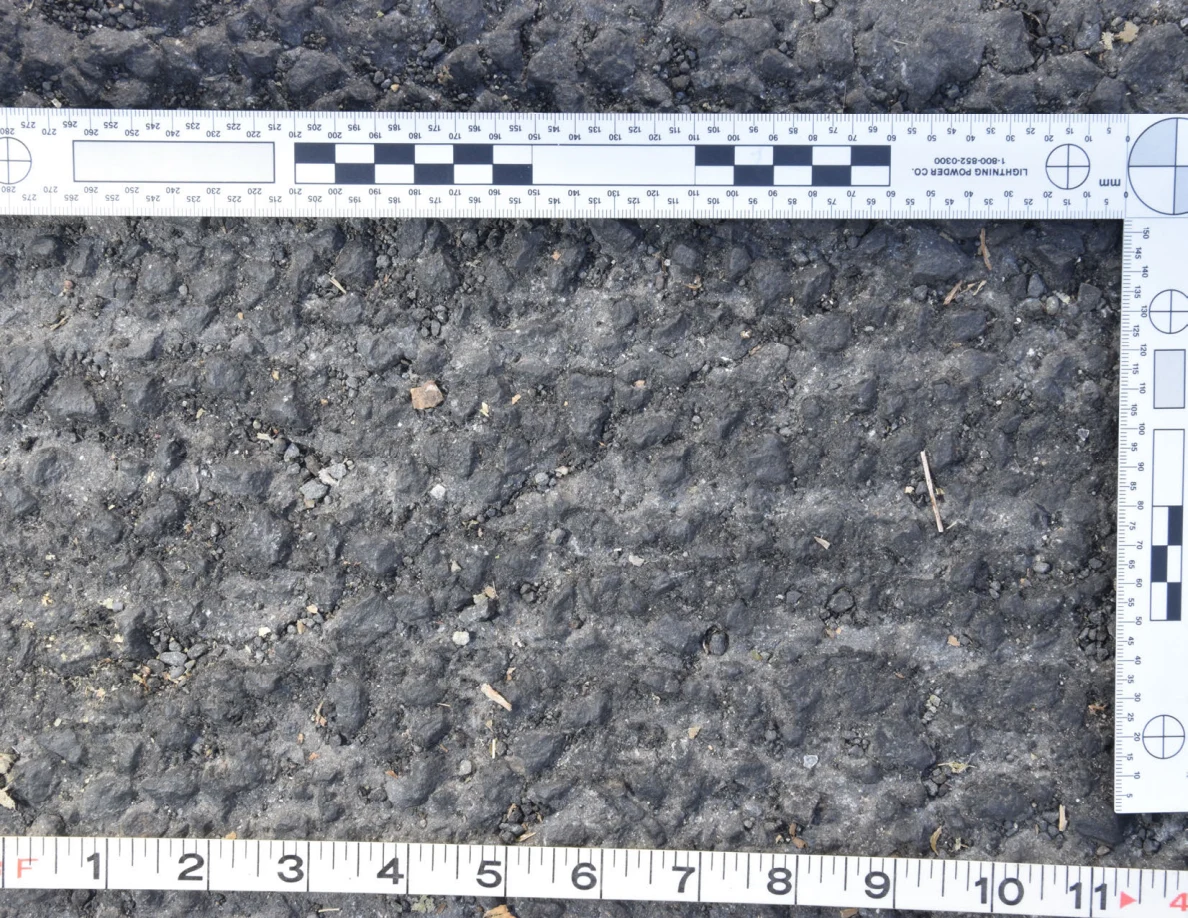 Close view of rough asphalt with small stones and thin plant fragments beside an L-shaped scale and measuring tape.