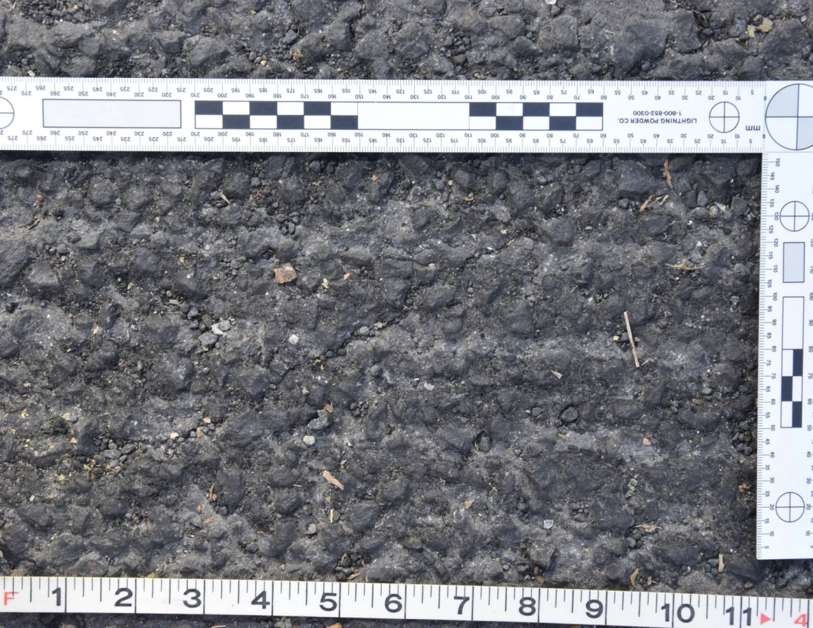 Close view of rough asphalt with small stones and thin plant fragments beside an L-shaped scale and measuring tape.
