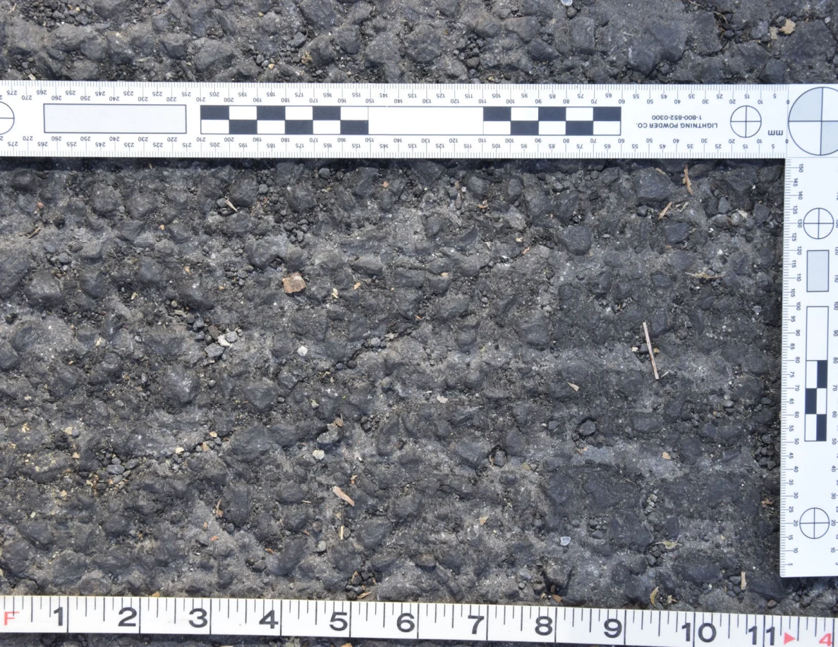 Close view of rough asphalt with small stones and thin plant fragments beside an L-shaped scale and measuring tape.
