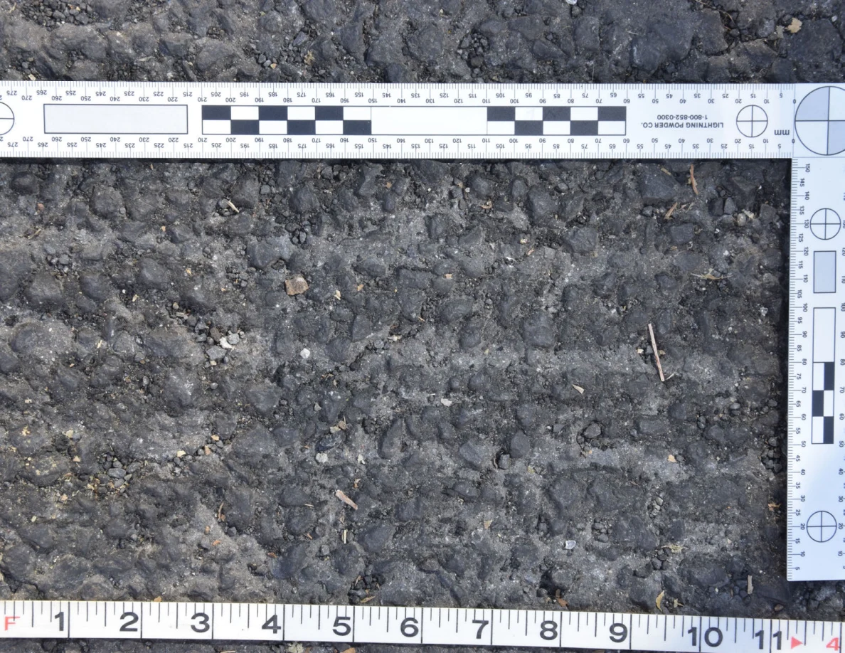 Close view of rough asphalt with small stones and thin plant fragments beside an L-shaped scale and measuring tape.