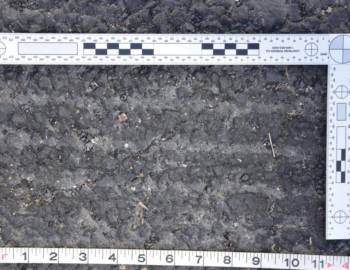 Close view of rough asphalt with small stones and thin plant fragments beside an L-shaped scale and measuring tape.