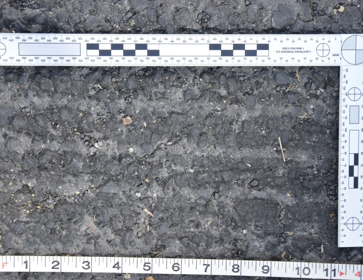 Close view of rough asphalt with small stones and thin plant fragments beside an L-shaped scale and measuring tape.