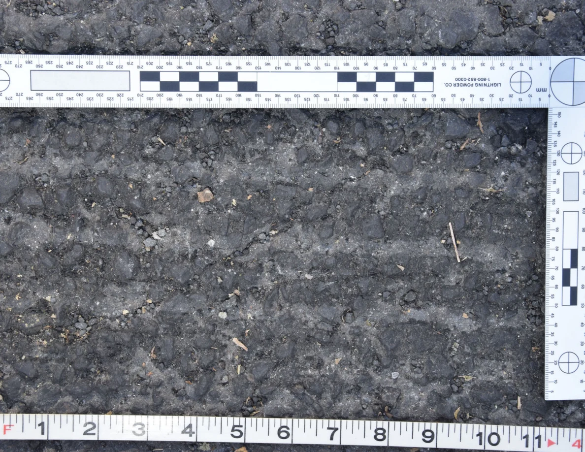Close view of rough asphalt with small stones and thin plant fragments beside an L-shaped scale and measuring tape.