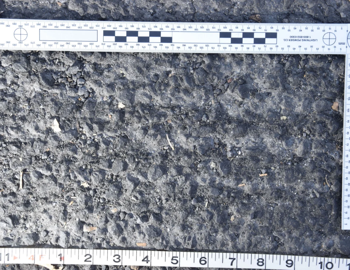 Uneven asphalt with pebbles and plant fragments, shown with an L-shaped photographic scale and measuring tape.