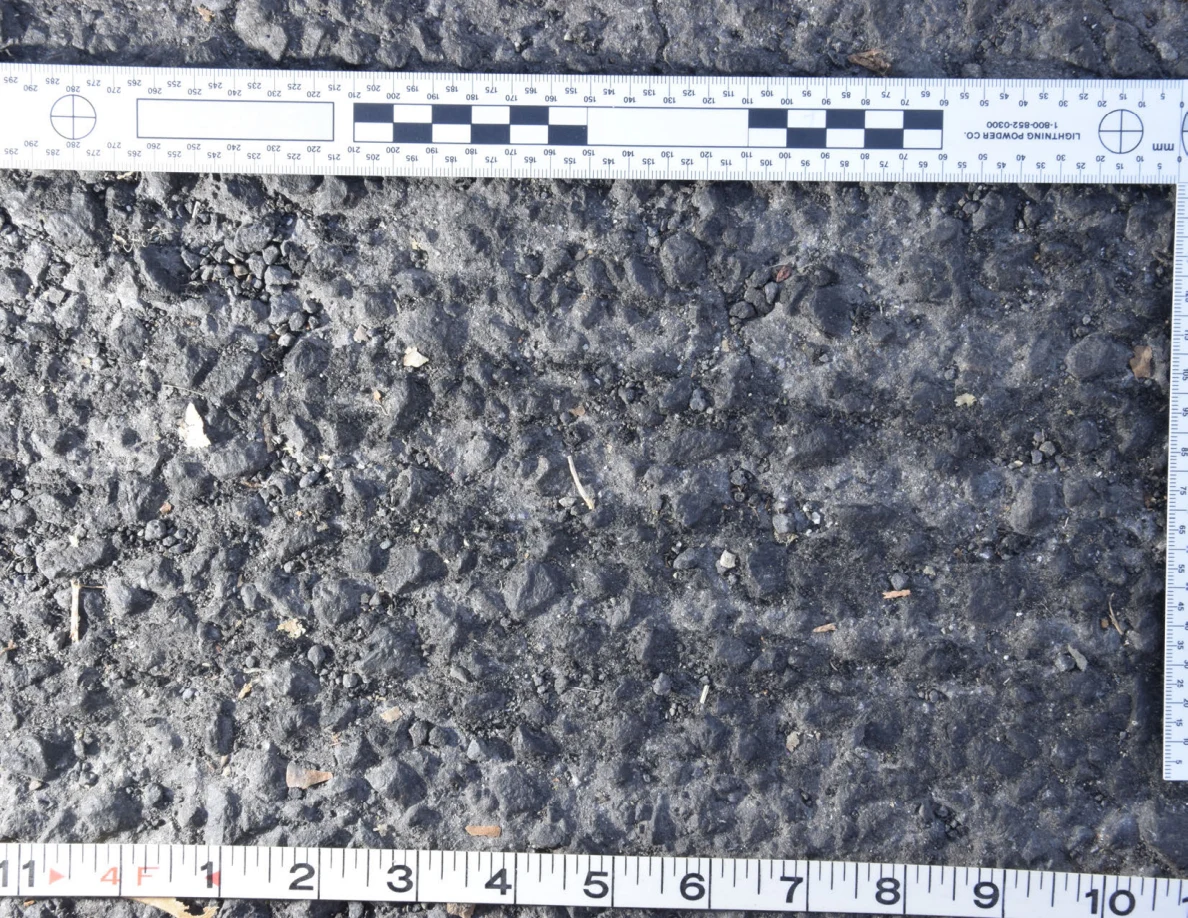 Uneven asphalt with pebbles and plant fragments, shown with an L-shaped photographic scale and measuring tape.