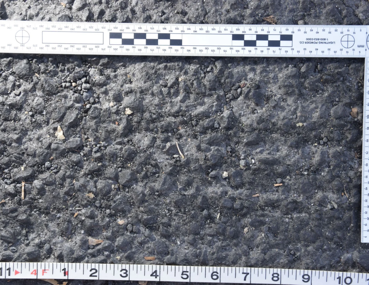 Uneven asphalt with pebbles and plant fragments, shown with an L-shaped photographic scale and measuring tape.