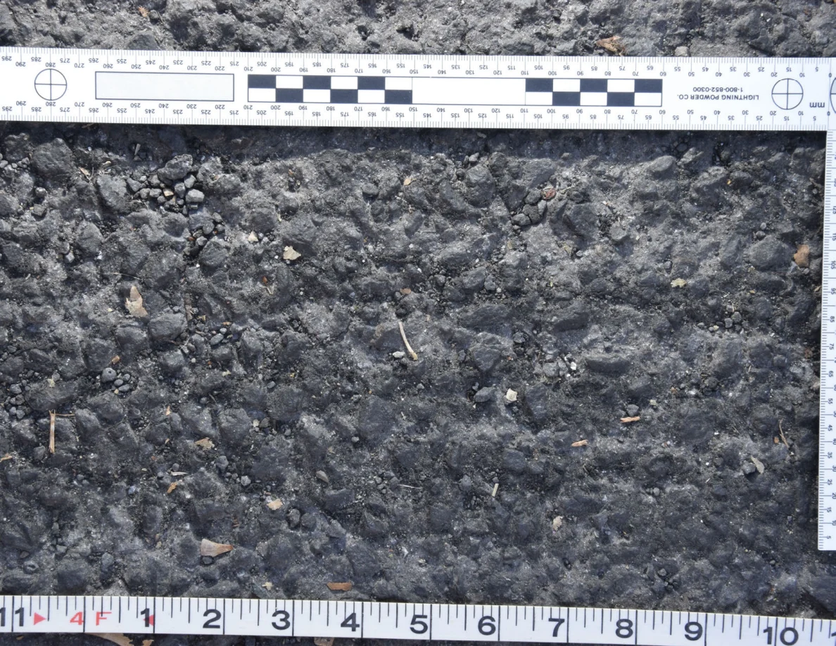 Uneven asphalt with pebbles and plant fragments, shown with an L-shaped photographic scale and measuring tape.