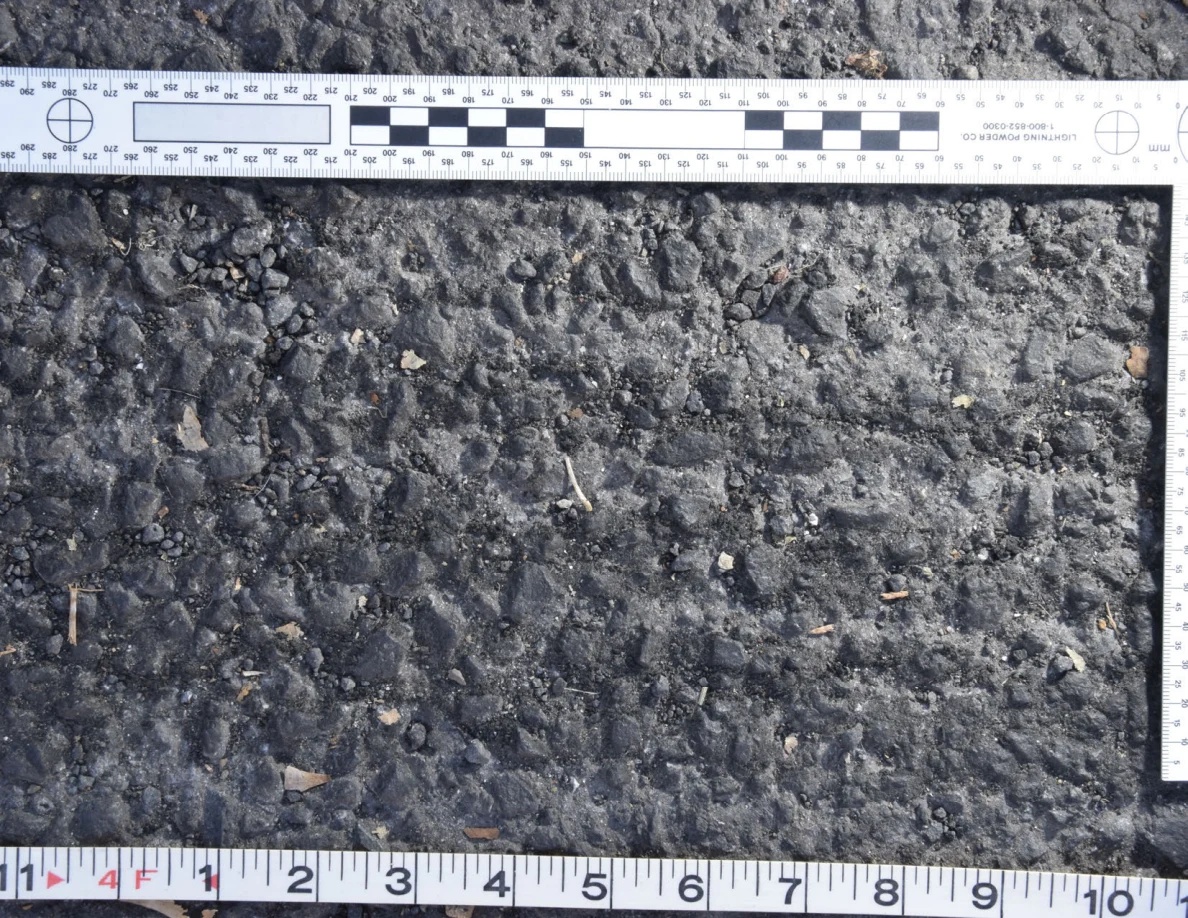 Close view of uneven asphalt with small stones and plant fragments, framed by an L-shaped scale and measuring tape.