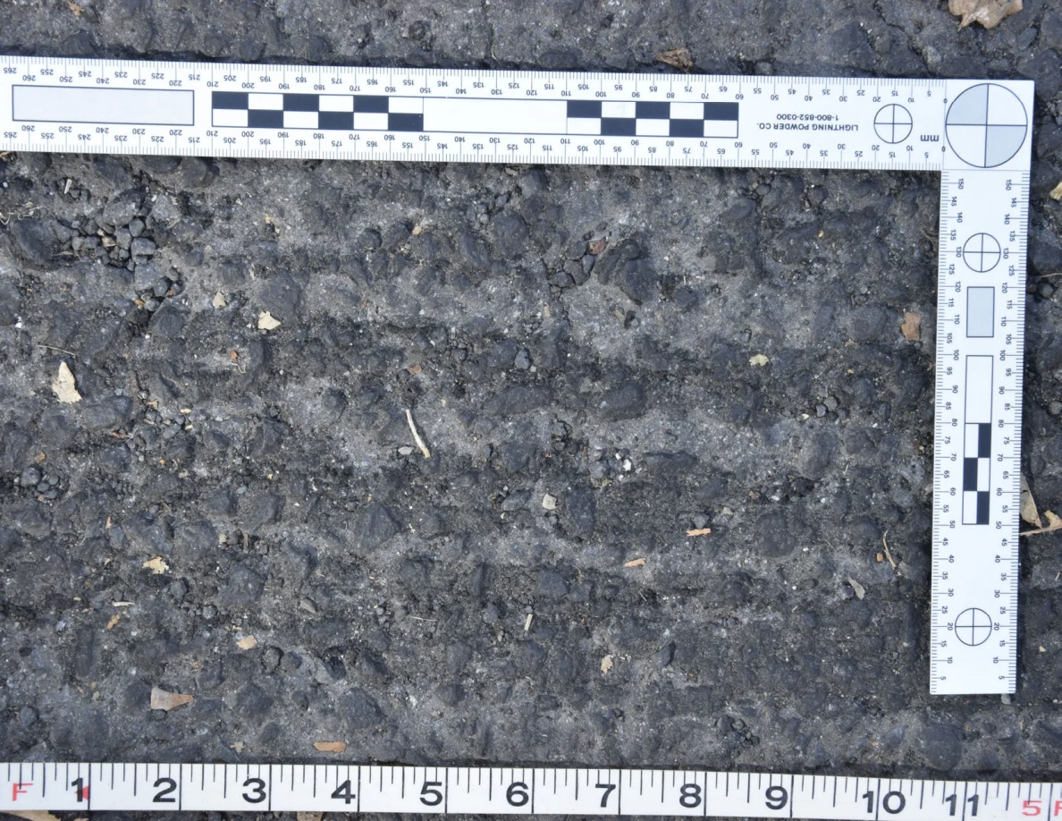 Close view of uneven asphalt with small stones and plant fragments, framed by an L-shaped scale and measuring tape.