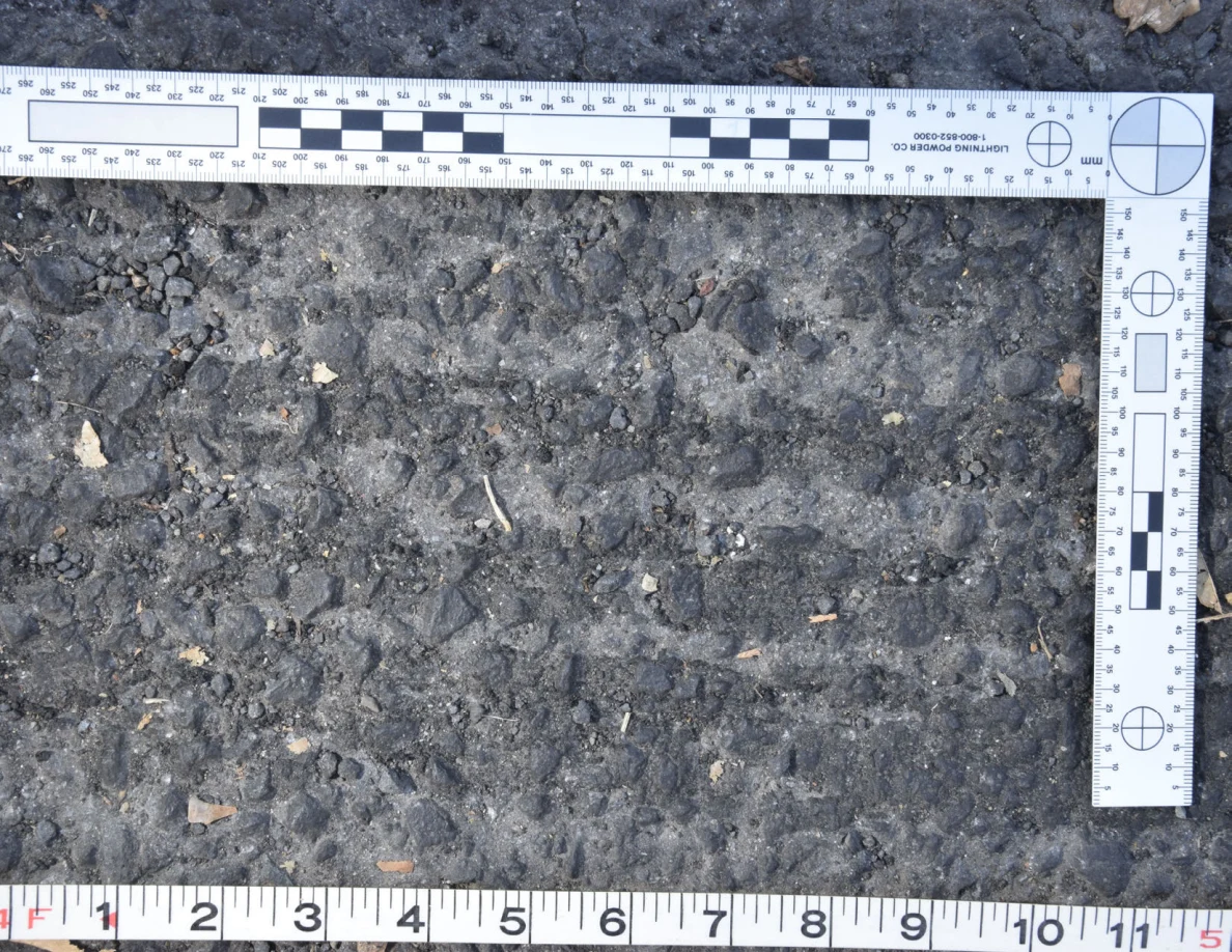 Close view of uneven asphalt with small stones and plant fragments, framed by an L-shaped scale and measuring tape.