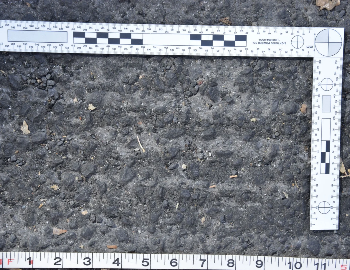 Close view of uneven asphalt with small stones and plant fragments, framed by an L-shaped scale and measuring tape.